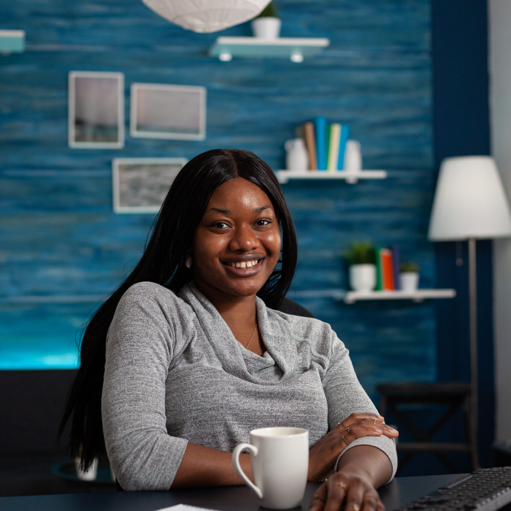 Portrait of cheerful person sitting at desk with computer, working from home on business project. Young woman looking at camera and using monitor with keyboard to do remote work.