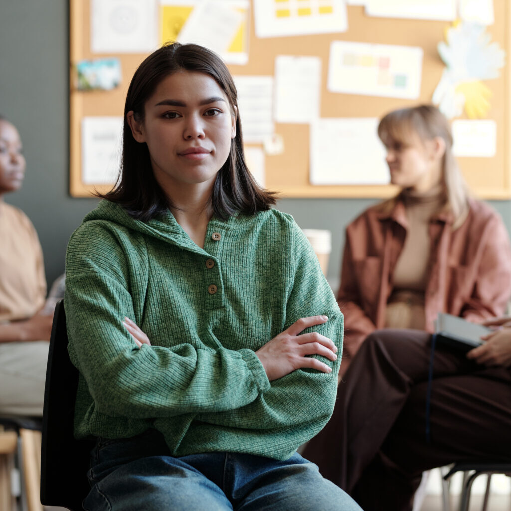 Young brunette woman in green casual pullover and blue jeans looking at camera while sitting against psychologist and patients having session